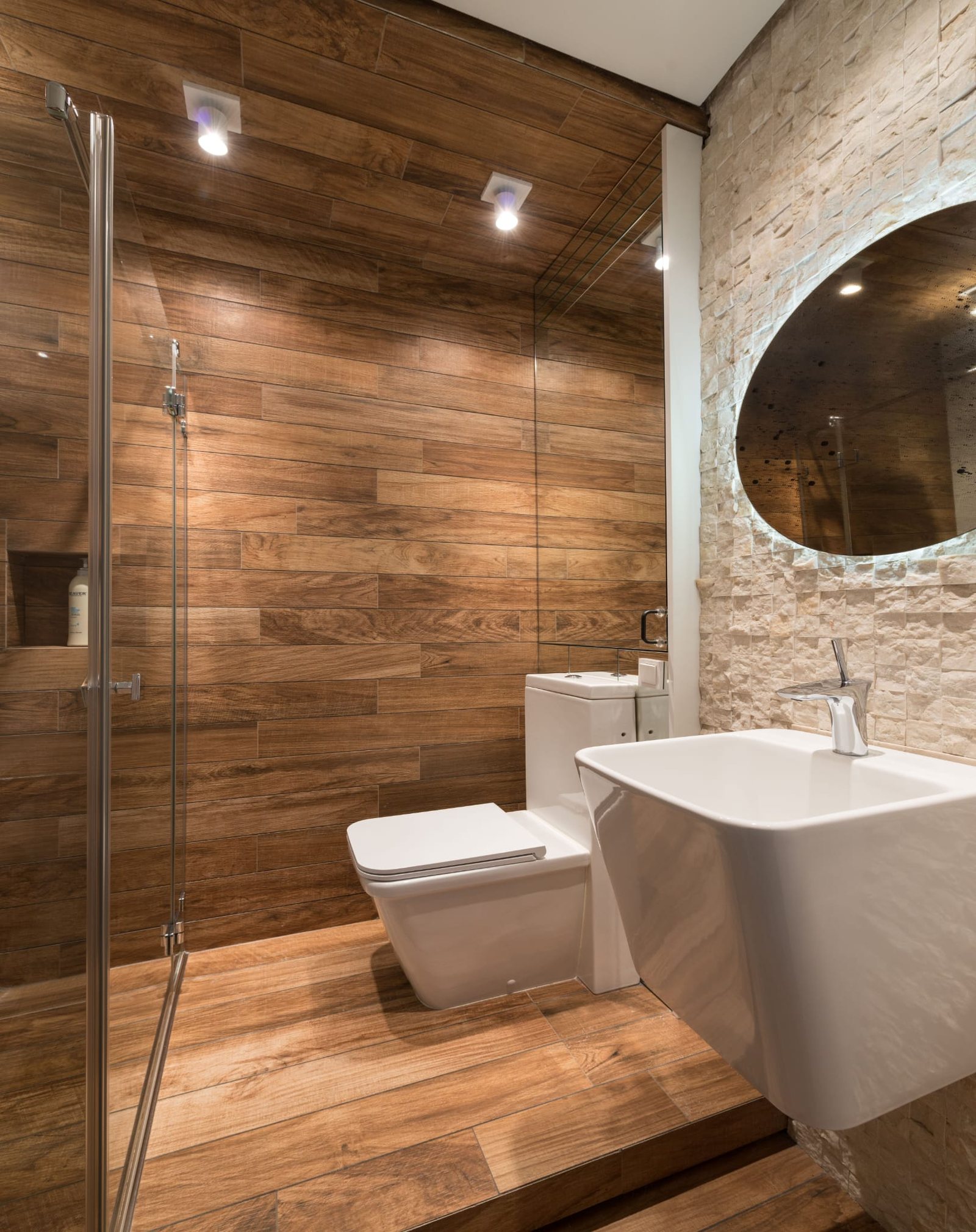 Ausgeführtes Badezimmer mit warmer Holzoptik, Glasdusche und einer Wand aus hellen Natursteinriemchen